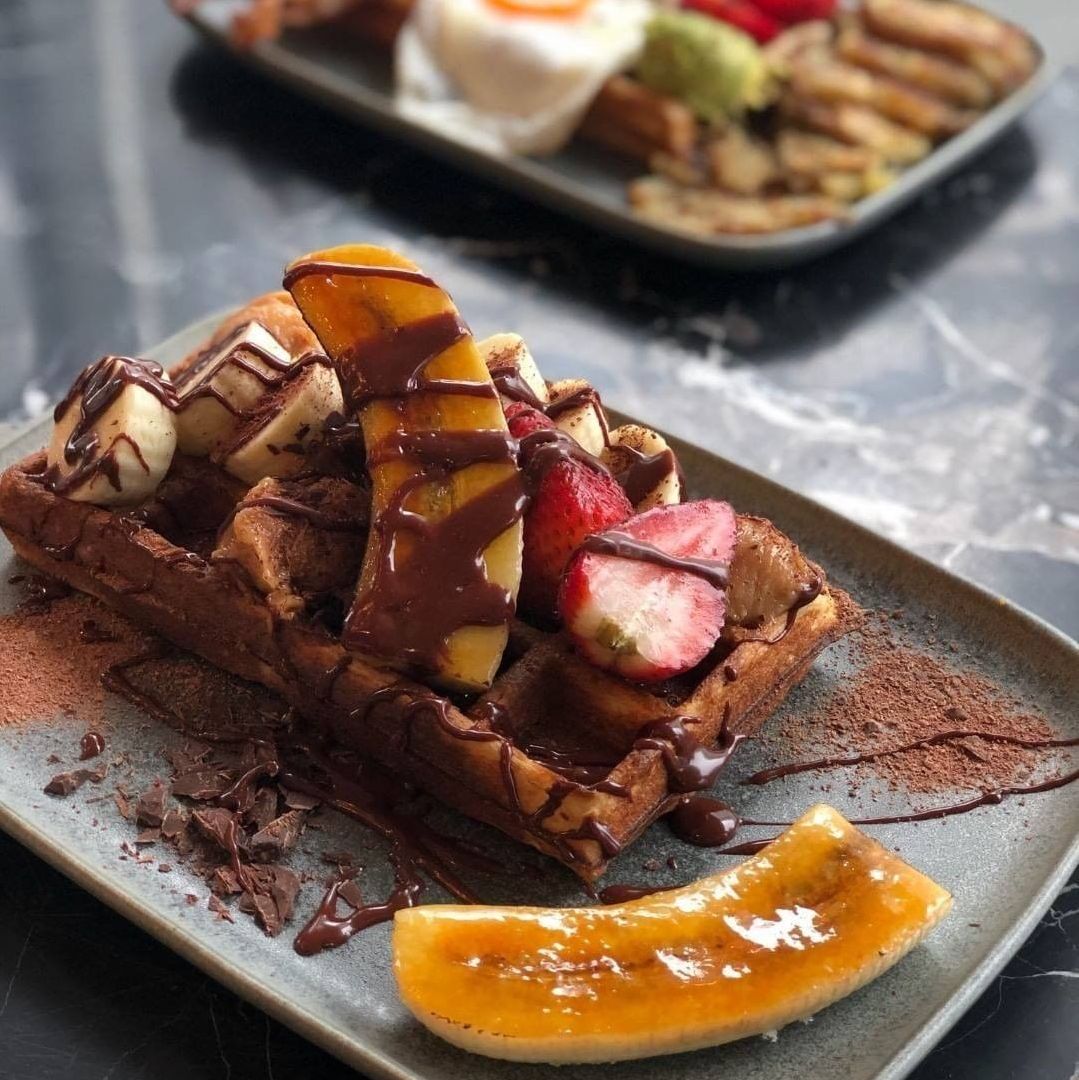 Assorted breakfast dishes including waffle topped with fruit and chocolate drizzle on a dark table